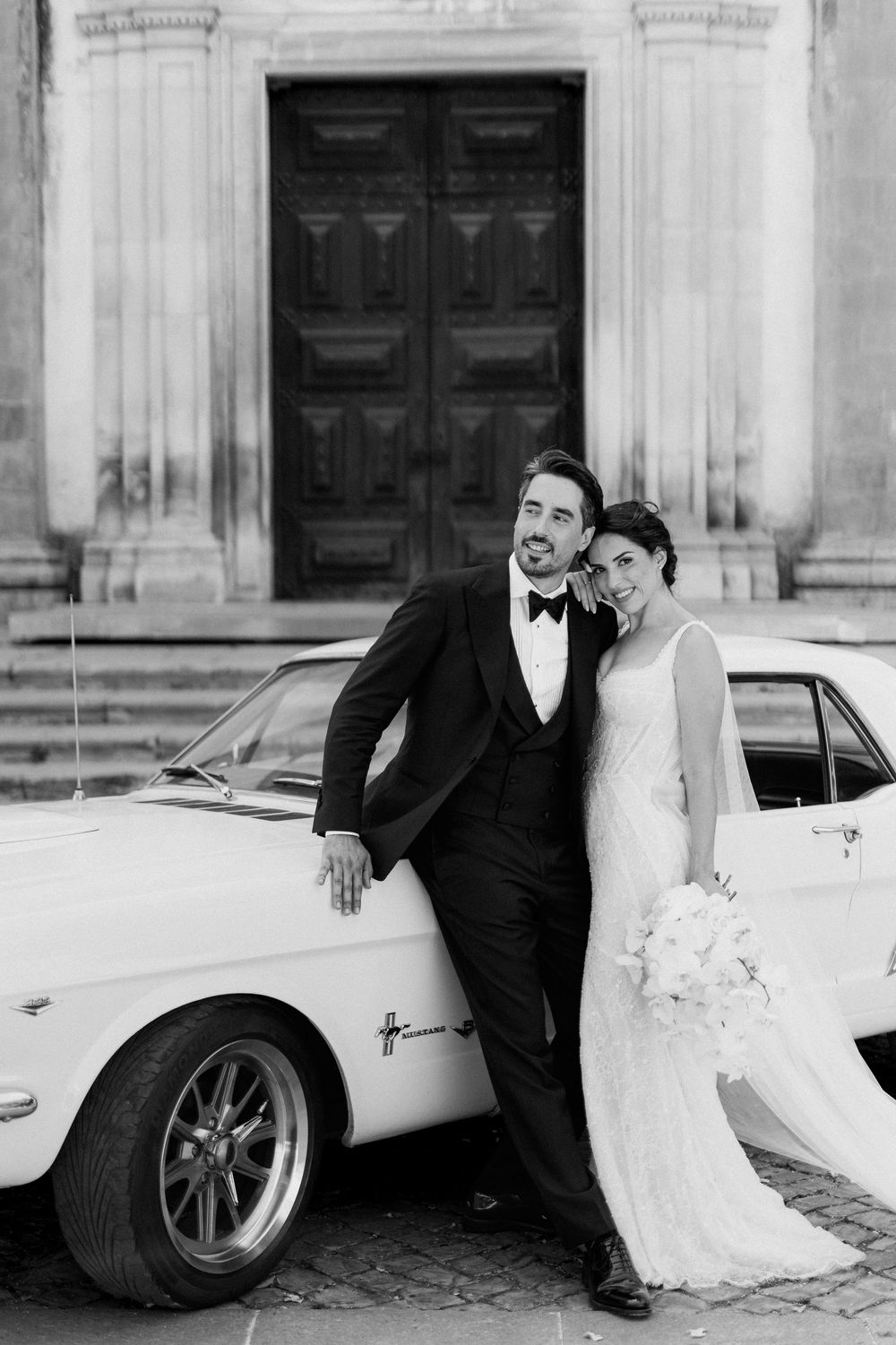 Black and white photo of a bride and groom leaning against a classic Mustang in front of a grand building.