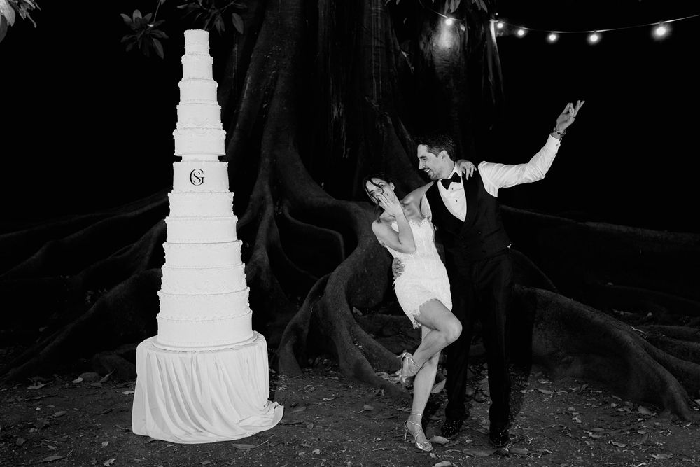 Black and white photo of bride and groom dancing beside tall elegant wedding cake outdoors at night.