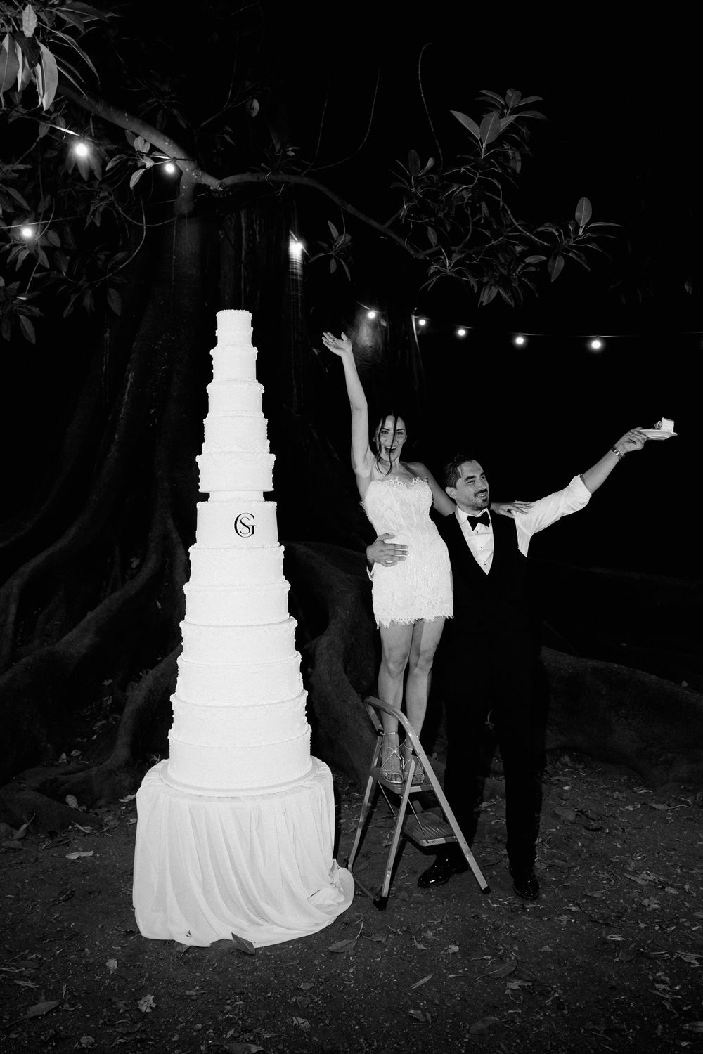 Bride and groom celebrating beside tall white wedding cake outdoors at night with string lights in black and white photo.