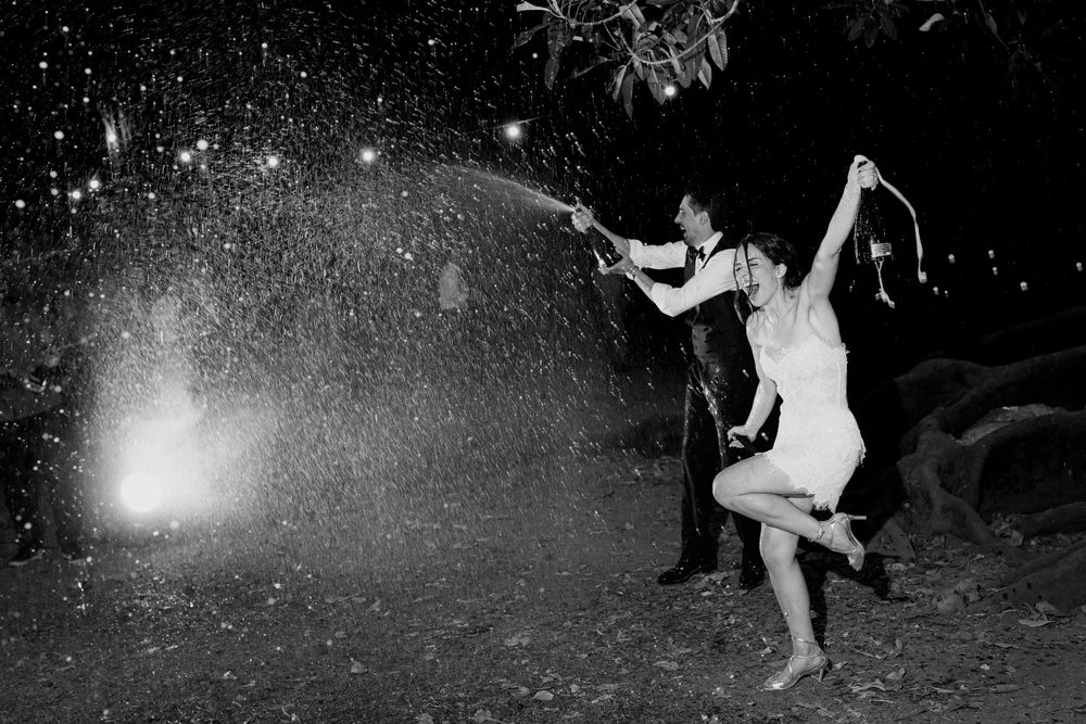 Black and white photo of a couple celebrating with champagne spray at a wedding reception outdoors at night.