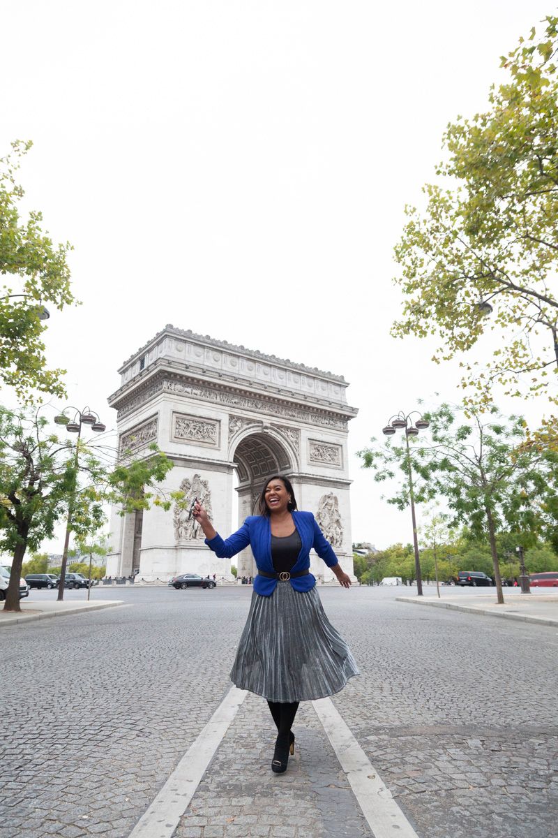 A person in a blue top and gray skirt poses joyfully in front of the Arc de Triomphe in Paris.