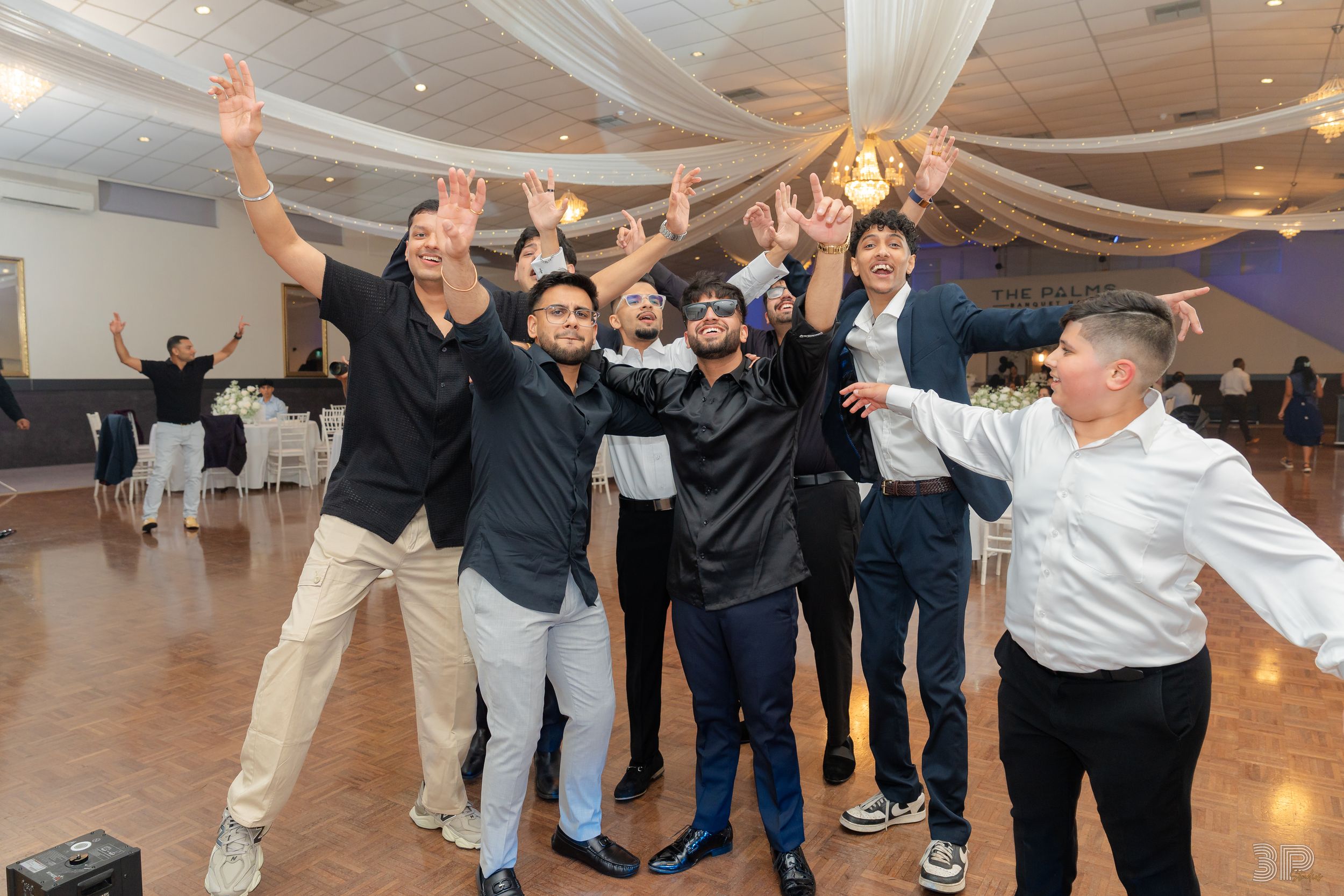 A group of celebratory guests cheer and raise their arms on a dance floor at an indoor reception venue.