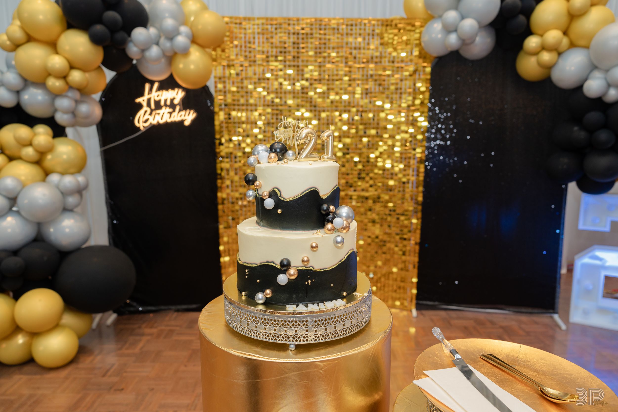 A three-tier black and white birthday cake with sparkly gold backdrop and balloon arch in gold, black and silver colors.