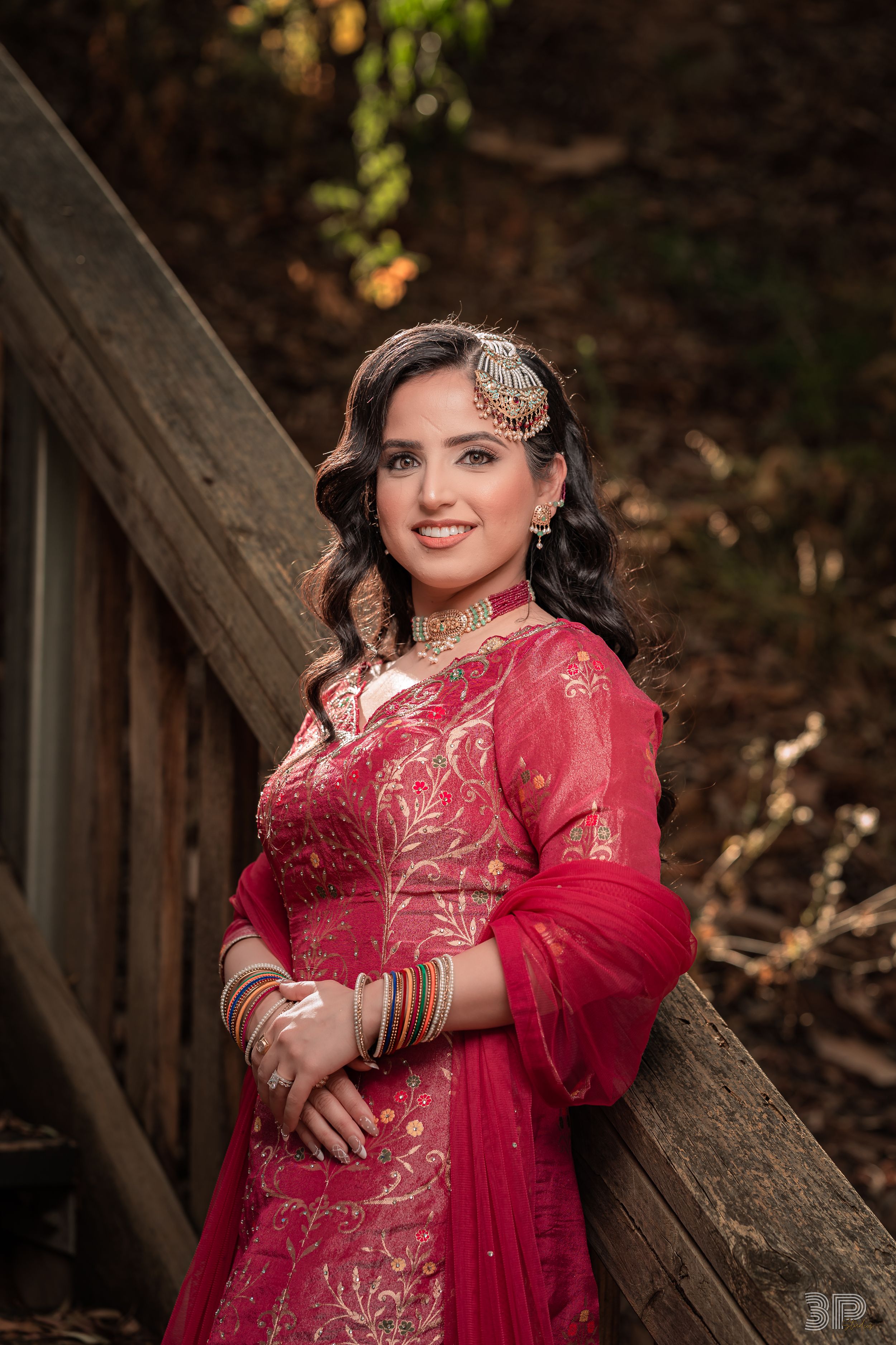 Traditional bride in a stunning red and gold embroidered dress poses elegantly against a wooden fence in an outdoor setting.