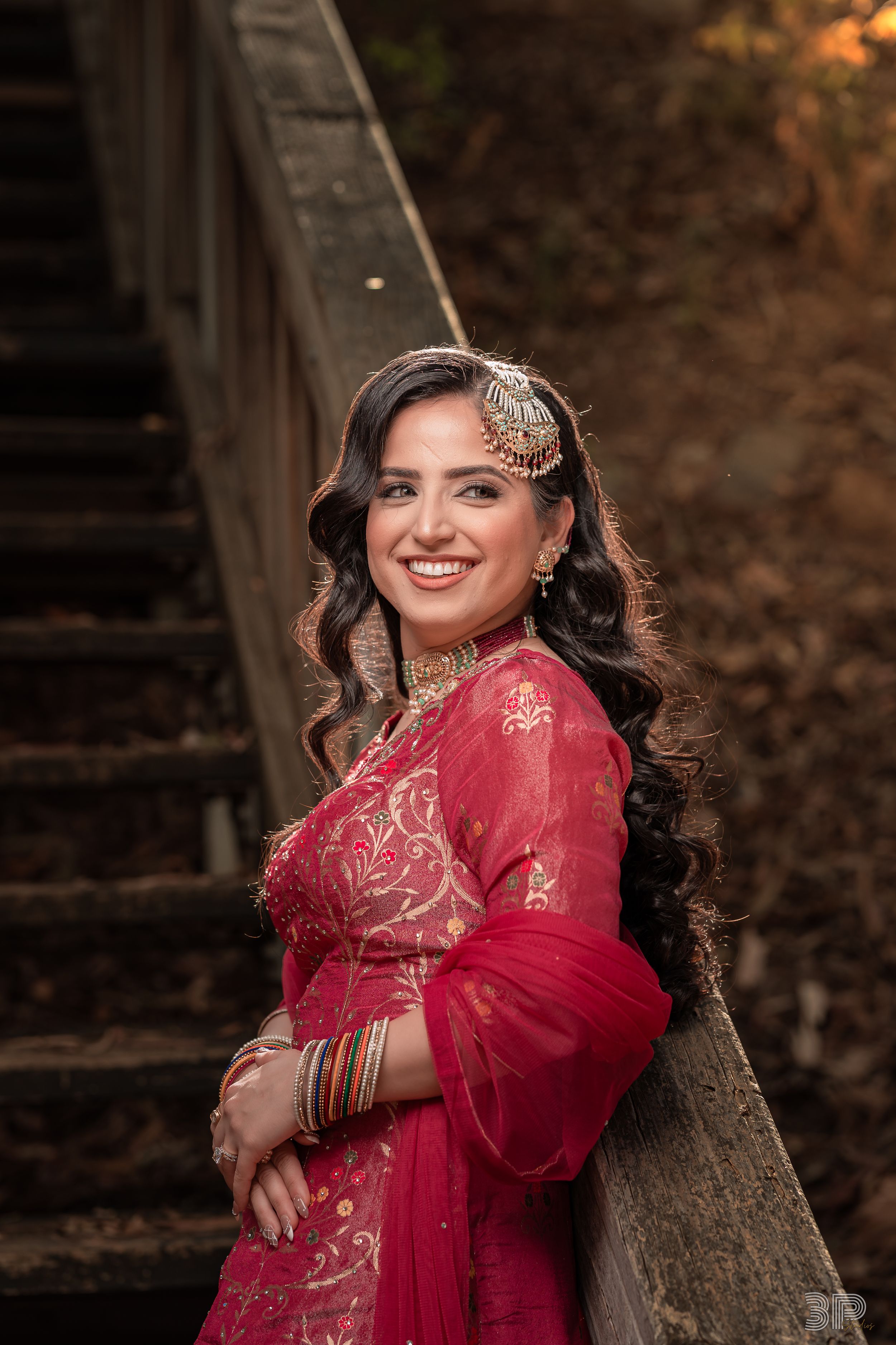 Person wearing a vibrant red traditional dress with gold embroidery poses elegantly on wooden stairs in an autumn setting.