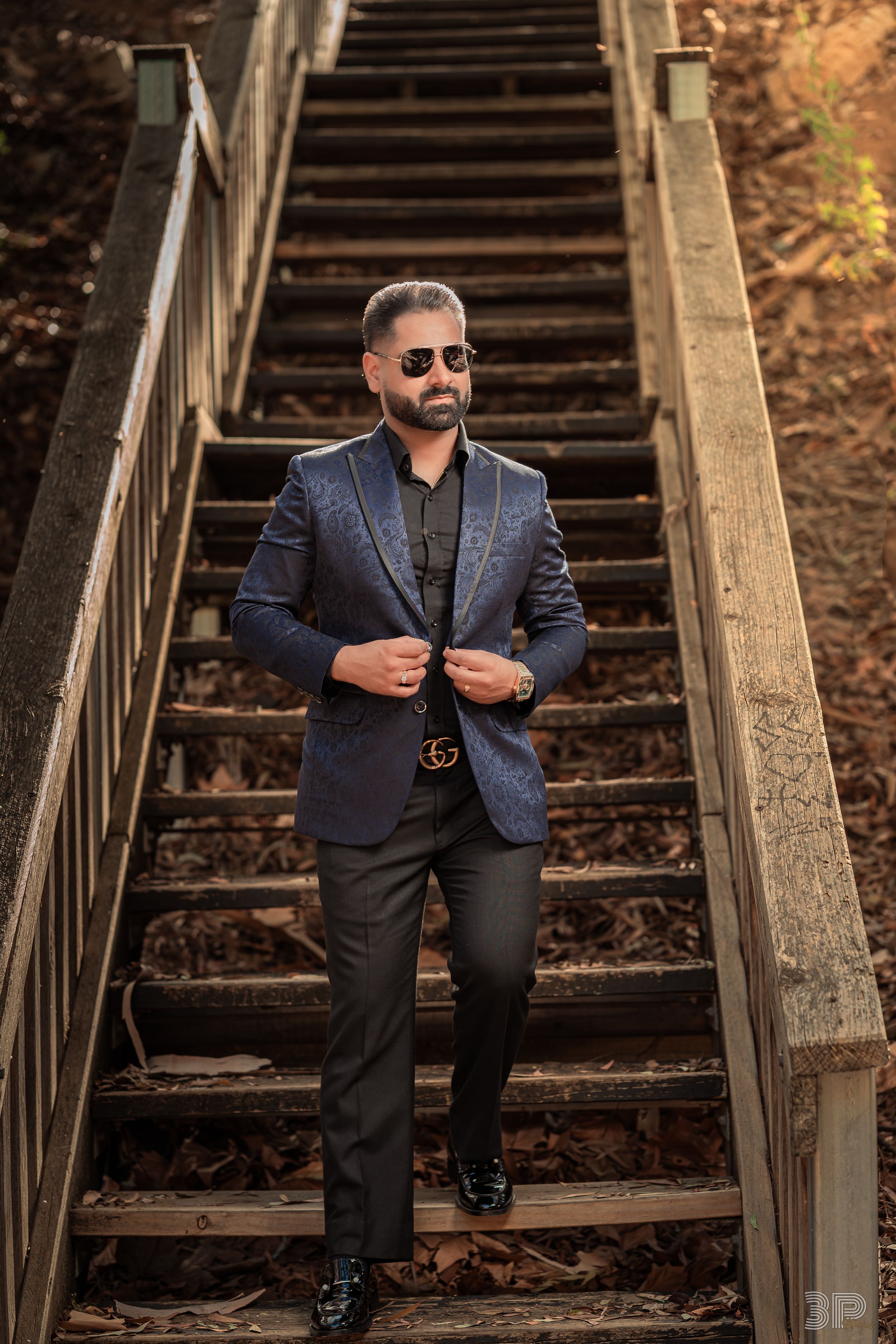 Person in dark suit and sunglasses poses confidently on rustic wooden stairs surrounded by fallen leaves.