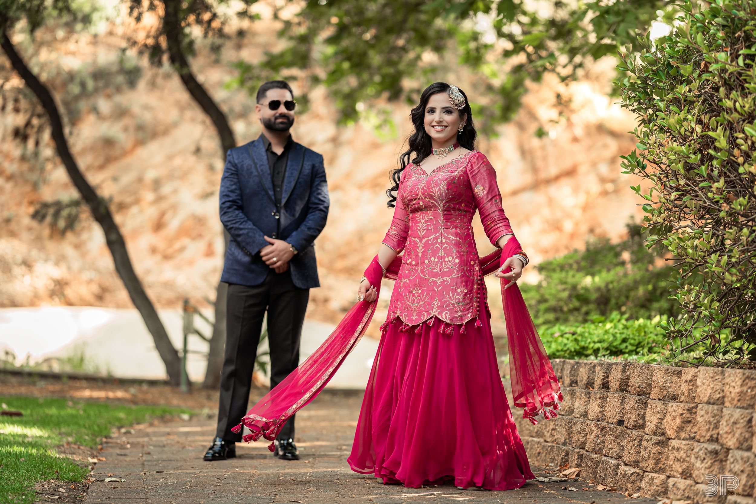 A couple poses outdoors in formal attire with a red lehenga and navy suit against a natural backdrop.