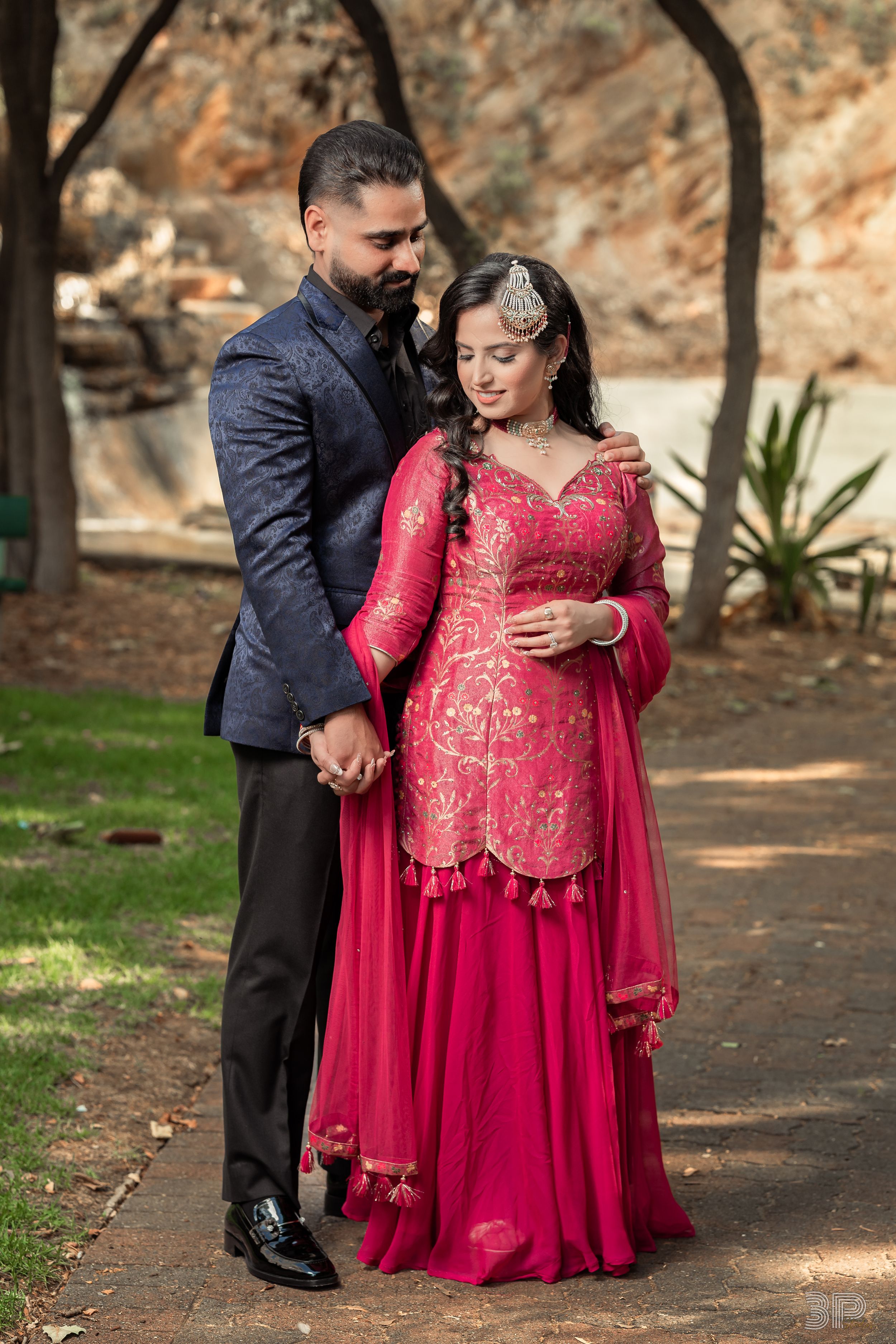 A couple poses for engagement photos in a park setting, with the woman wearing an elegant red and gold dress.