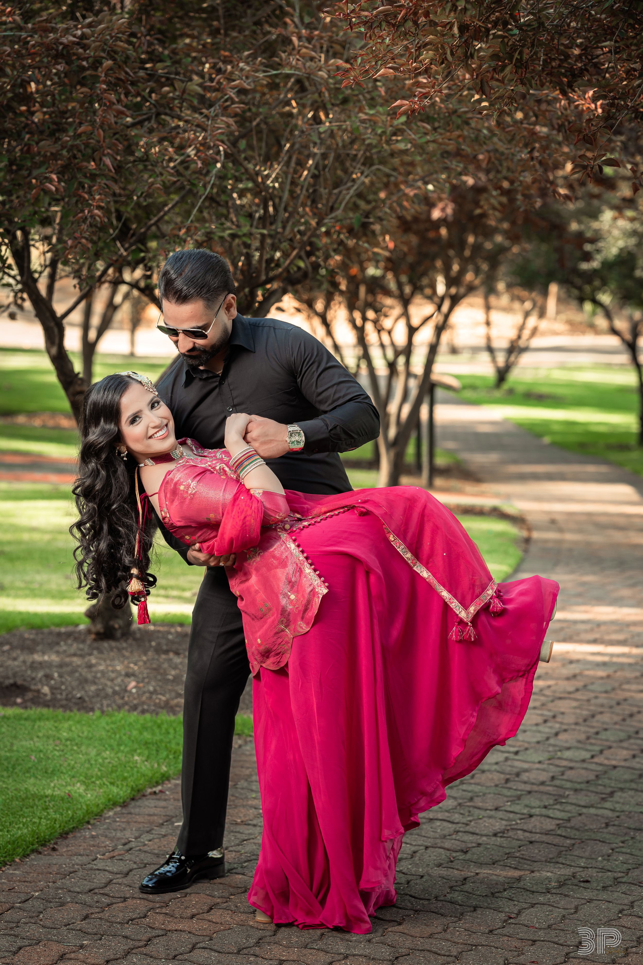 A romantic couple poses on a brick pathway, with her in a flowing red gown and him in a black suit.