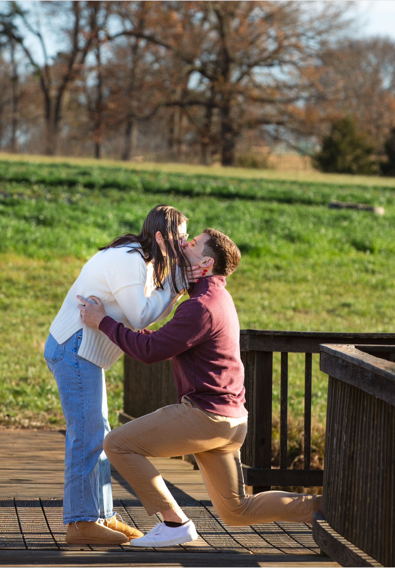 A couple shares an intimate moment on a wooden deck overlooking a grassy hill on a sunny autumn day for their surprise proposal
