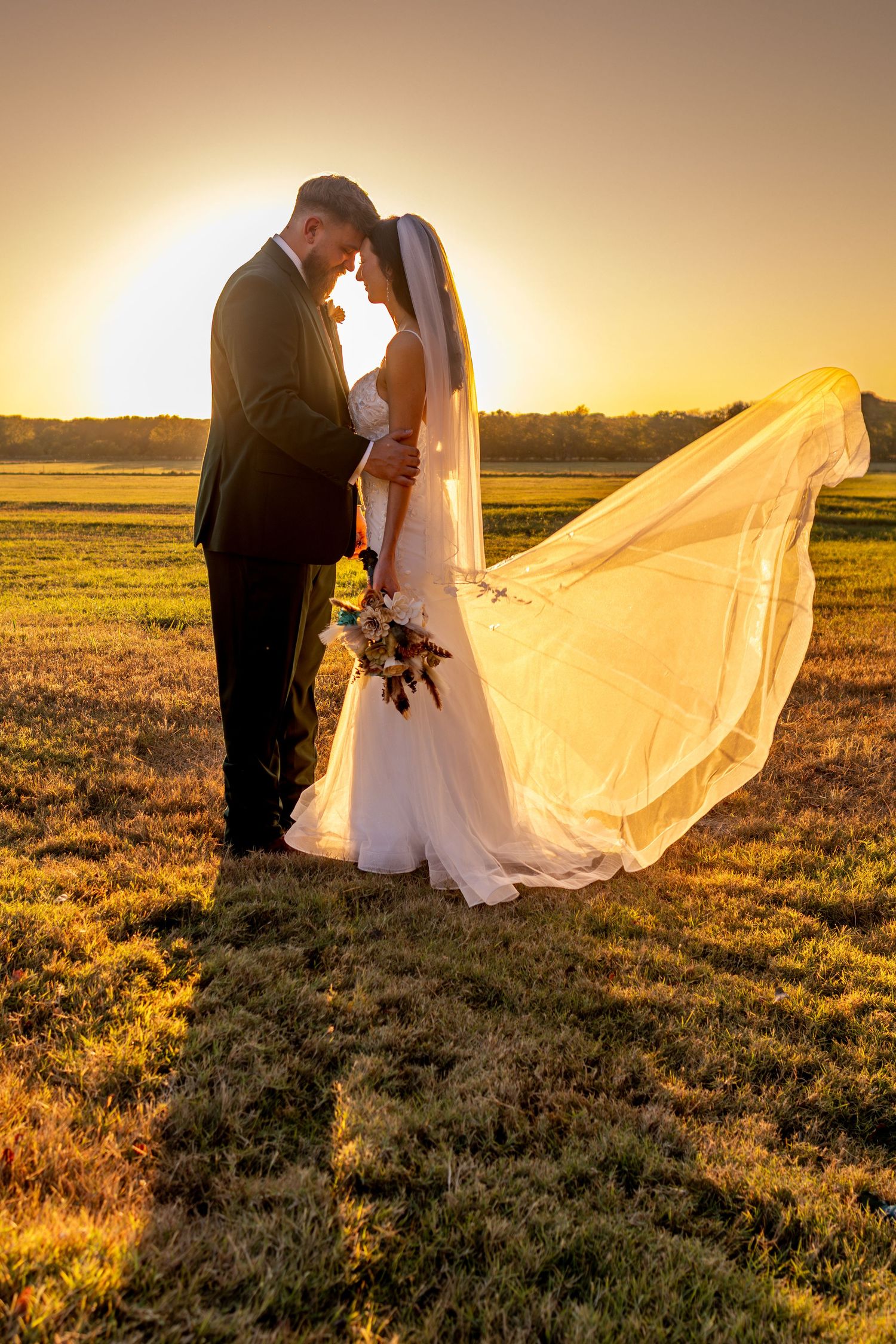 A wedding couple stands in a golden field at sunset as the bride's veil flows dramatically in the wind.
