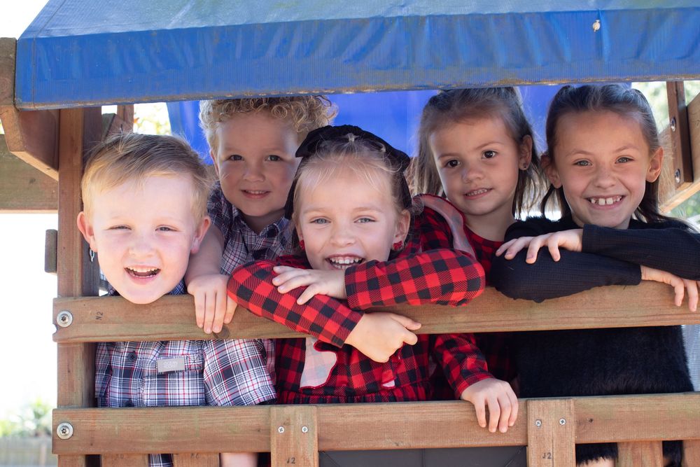 Group of children peering over a wooden railing with bright smiles and festive red plaid clothing.