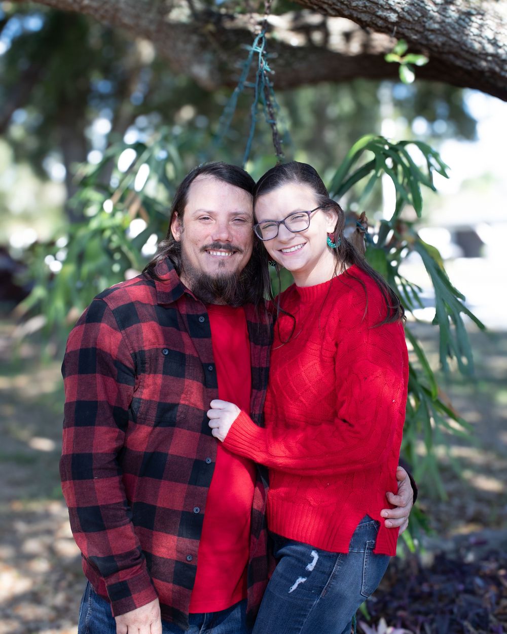 A couple in matching red and black buffalo plaid attire embraces outdoors under tree branches.