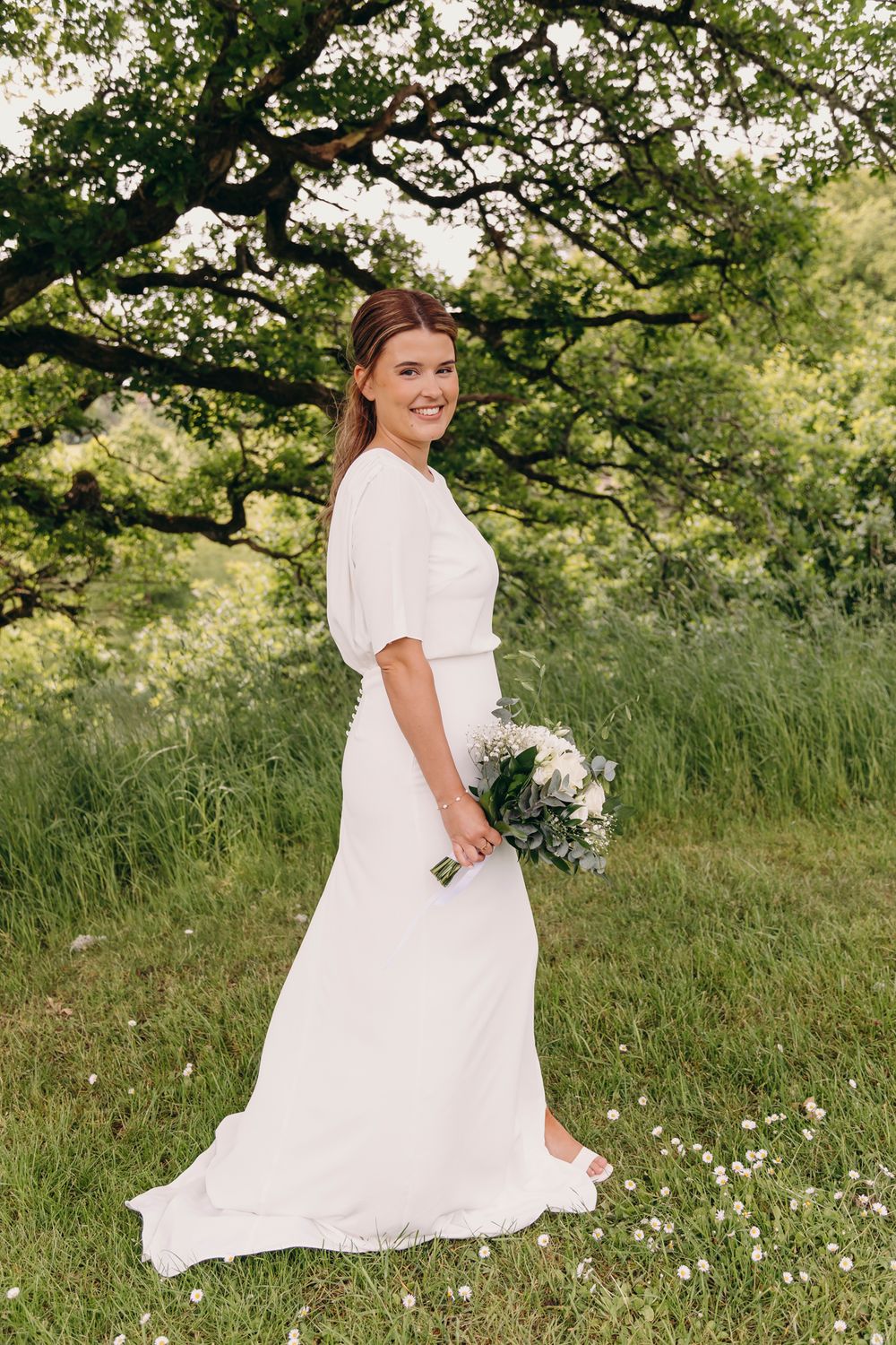 Bride in a flowing white dress poses in a sunlit meadow with wildflowers and trees in the background.