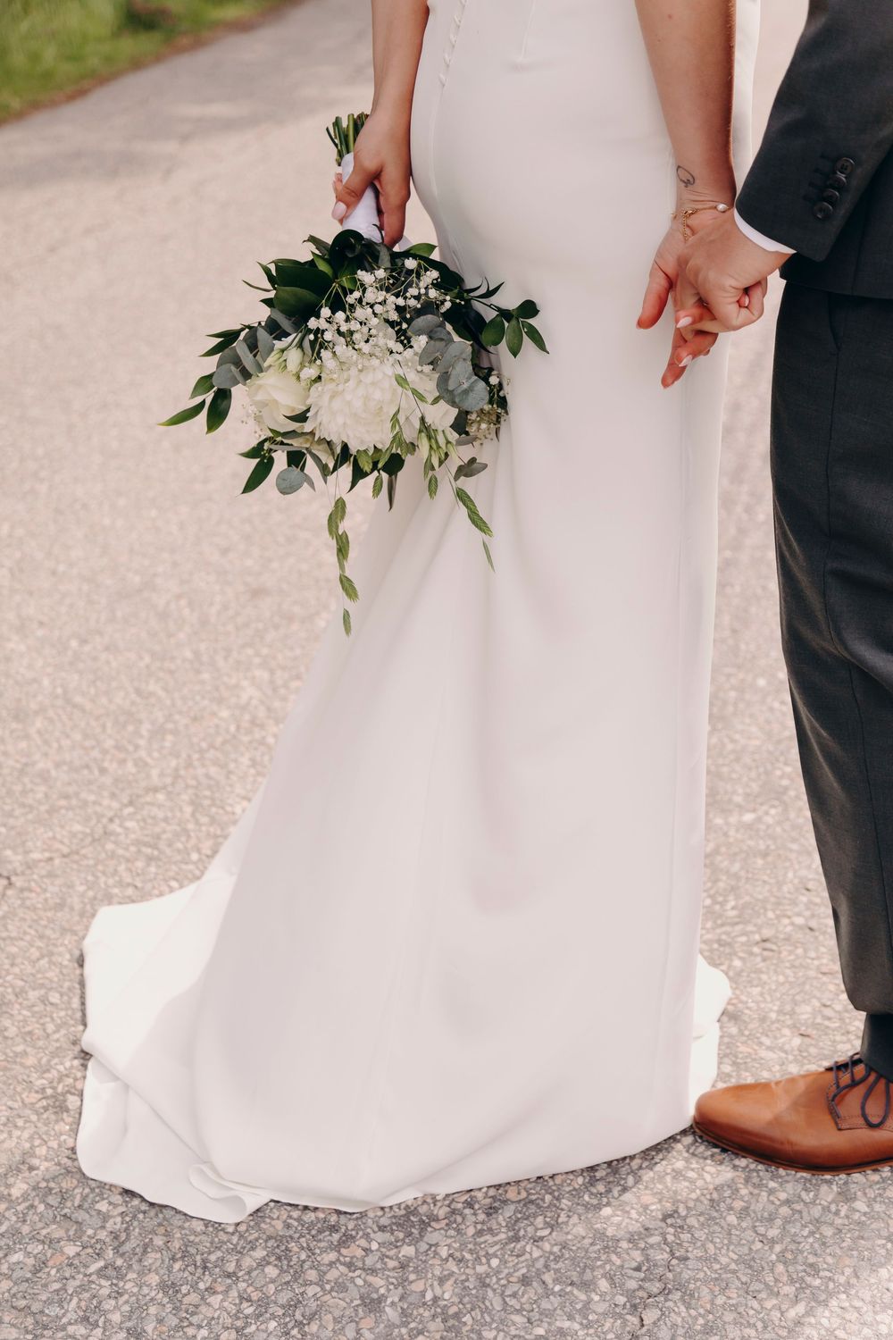 Close-up view of a white wedding dress hem and brown leather shoes on a gravel path with delicate greenery.