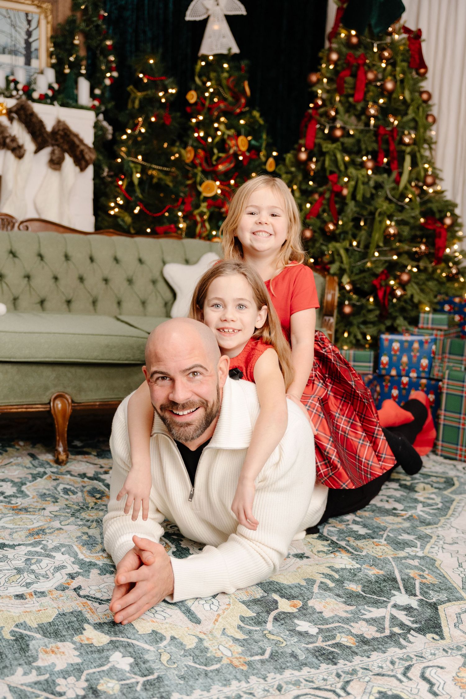 Family members lay together on a patterned carpet in front of decorated Christmas trees during holiday photoshoot.