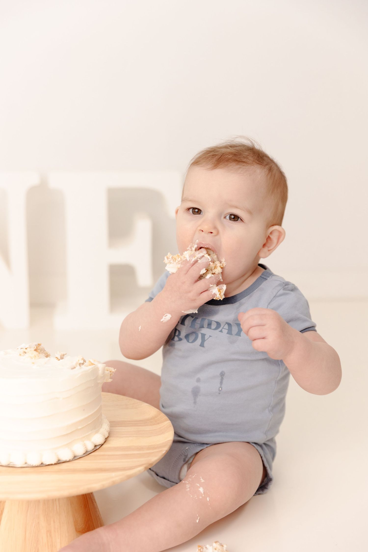 Baby in gray onesie sits on white floor enjoying birthday cake smash photo session.