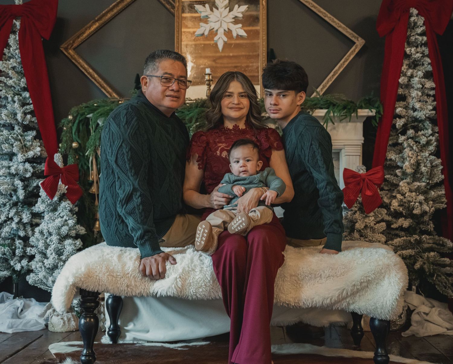 A family poses together in front of snow-covered Christmas trees in a cozy indoor holiday setting.