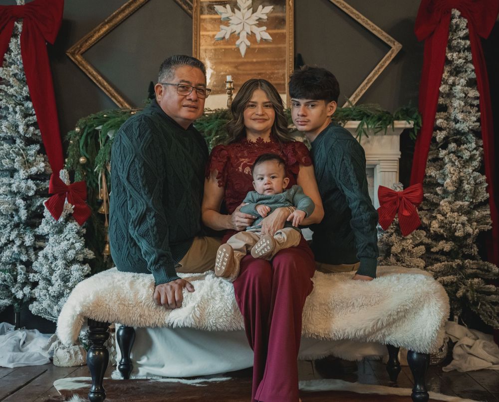 A family poses together in front of snow-covered Christmas trees in a cozy indoor holiday setting.