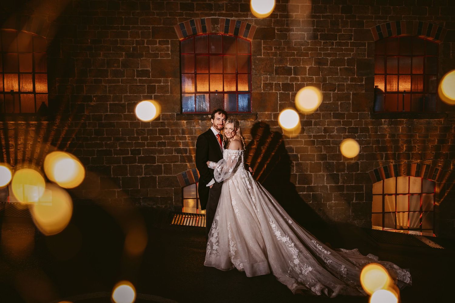 A romantic wedding couple dances in a rustic brick venue illuminated by warm glowing string lights and lanterns.