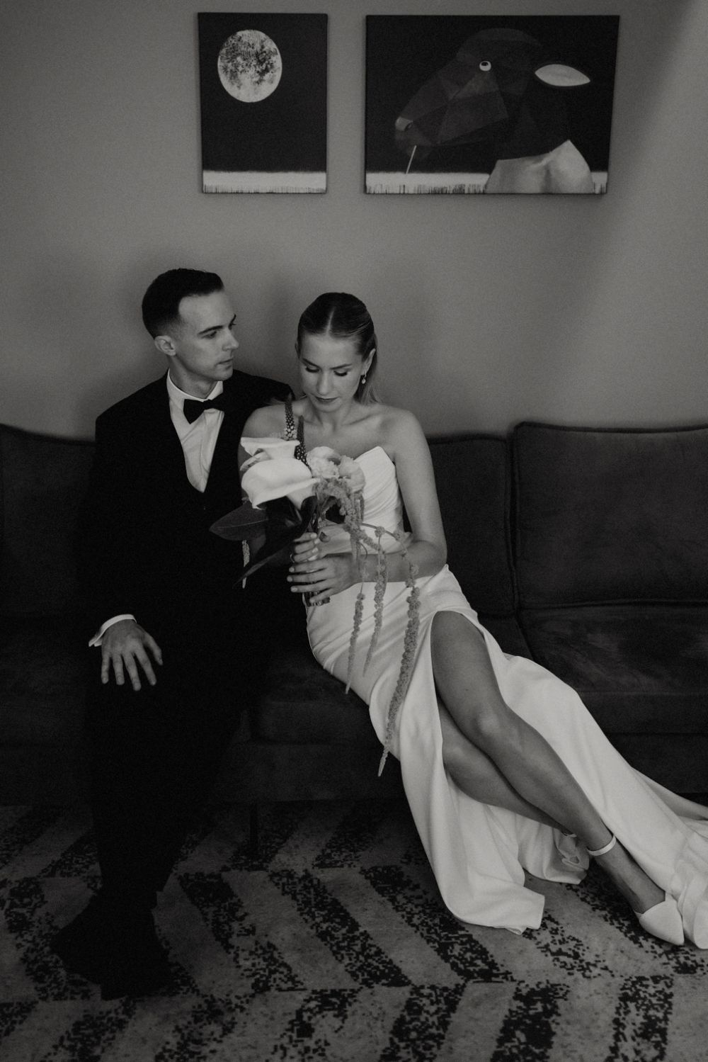 Couple in formal wear pose on couch beneath artistic moon paintings.