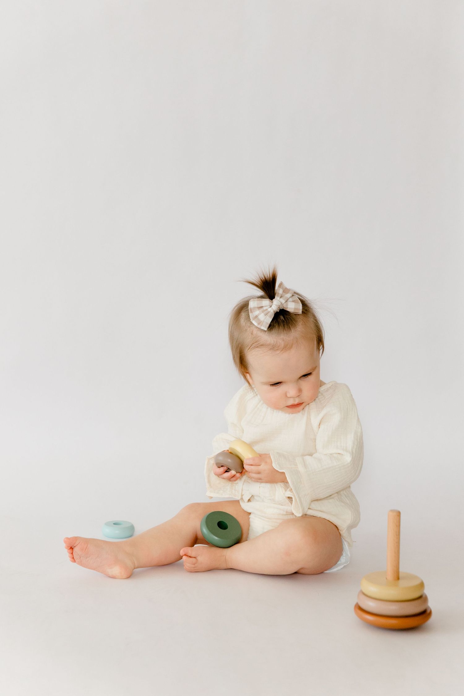 Adorable baby girl in cream bodysuit with bow sitting on white backdrop playing with stacking toy.