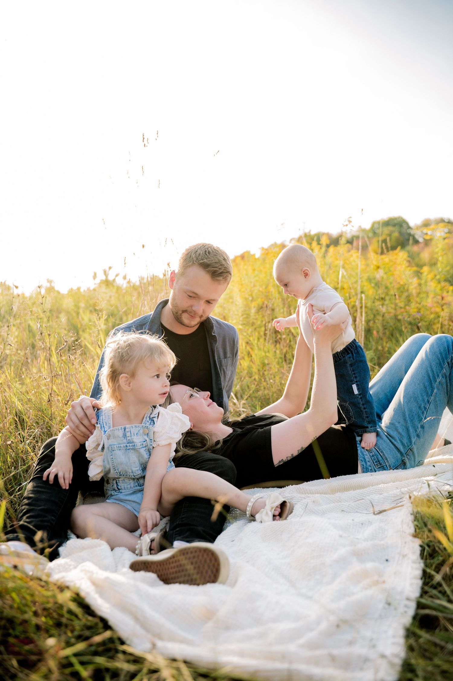 Family relaxing together on white blanket in golden sunlit meadow during outdoor photoshoot.