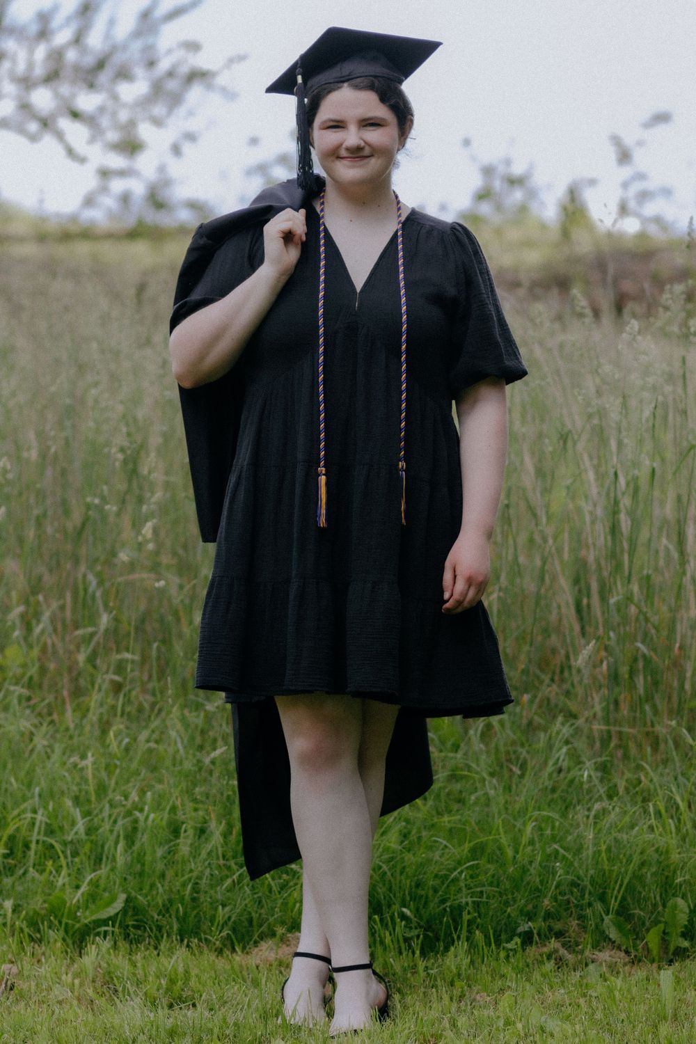 Graduate posing in traditional black cap and gown against a grassy field background.