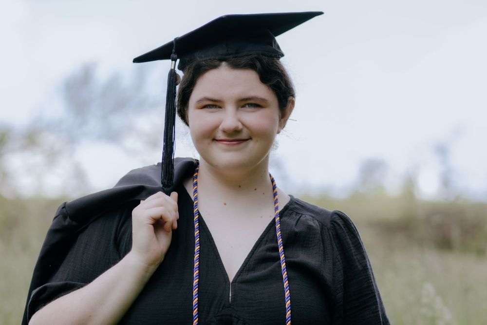 Graduate in academic regalia smiles while posing in outdoor setting.