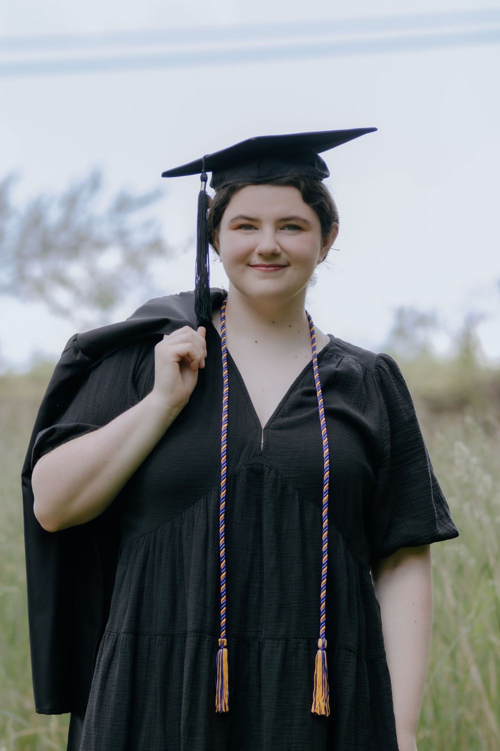 Graduate in black academic dress poses with graduation cap in meadow setting.