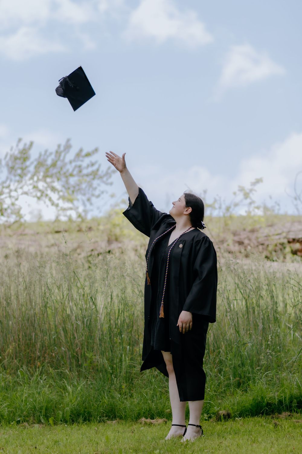 Graduate reaching up to catch mortarboard cap against blue sky and grass.