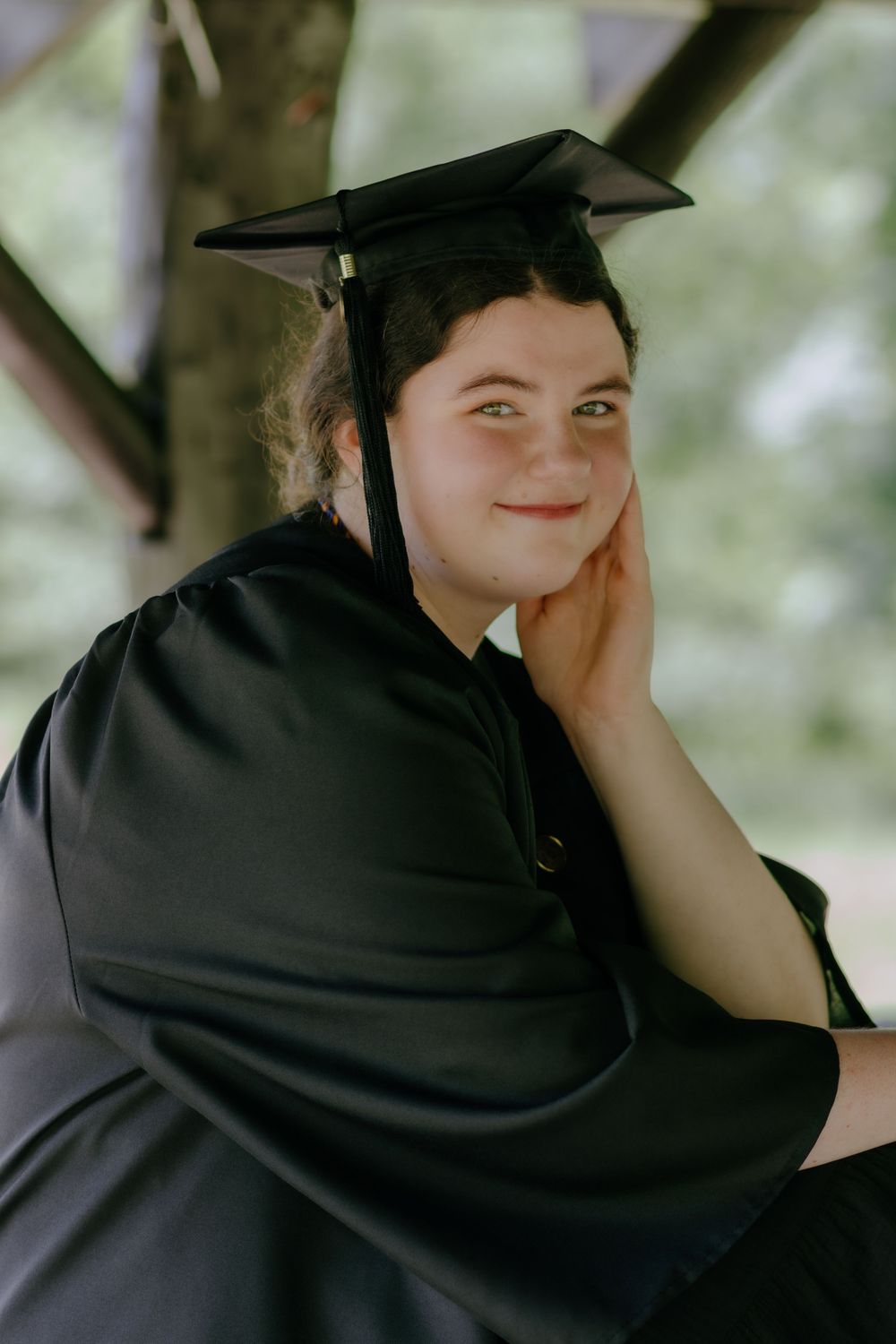 Graduate poses in traditional black graduation attire while sitting outdoors.