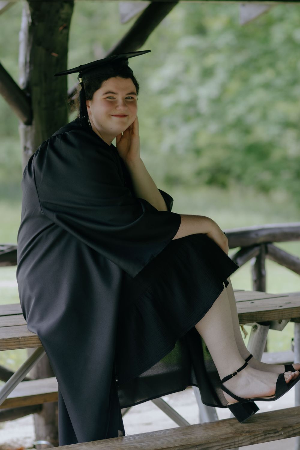 Graduate in black cap and gown sits on wooden bench during outdoor graduation photoshoot.