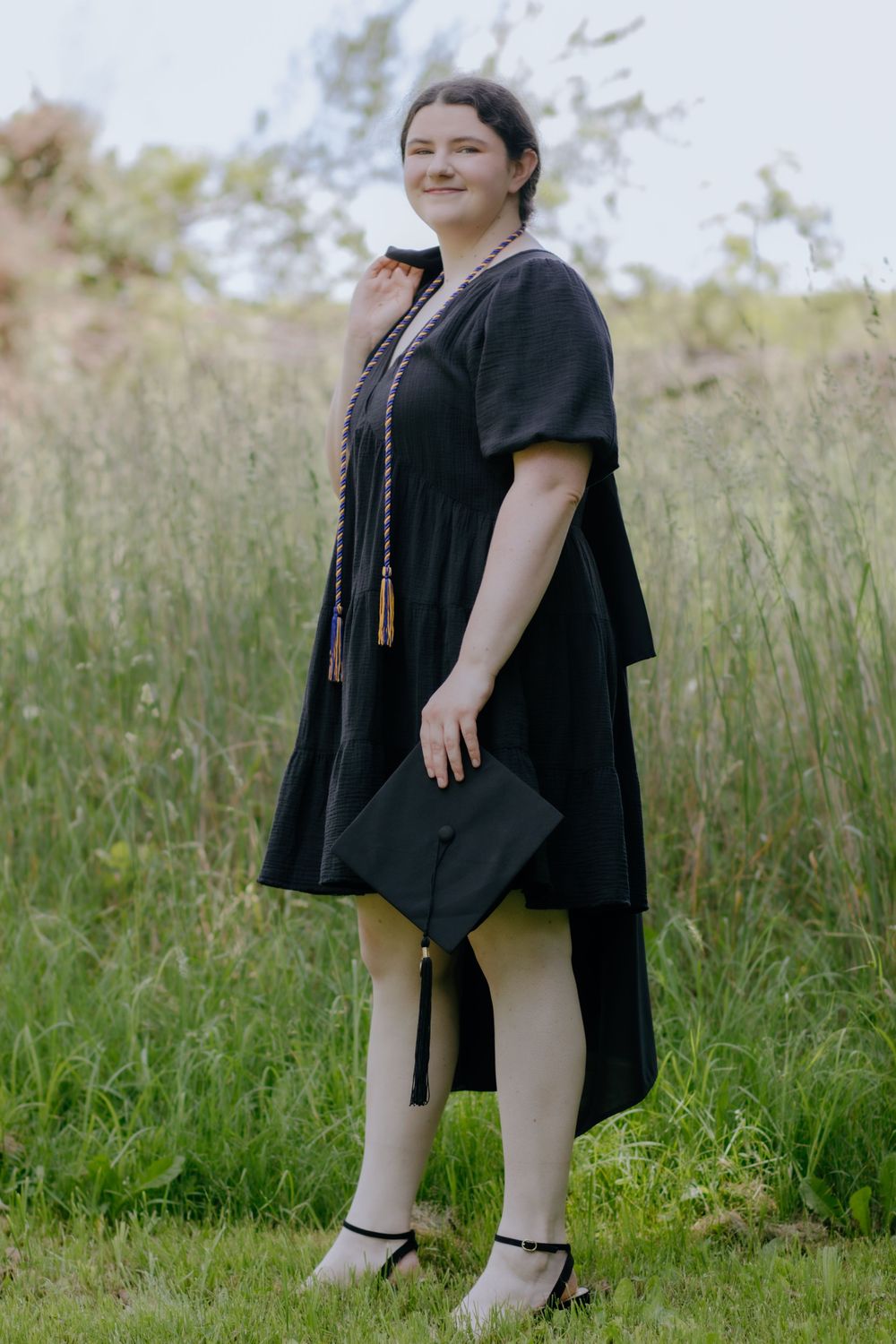 Graduate stands in grassy field wearing black graduation attire and holding diploma.