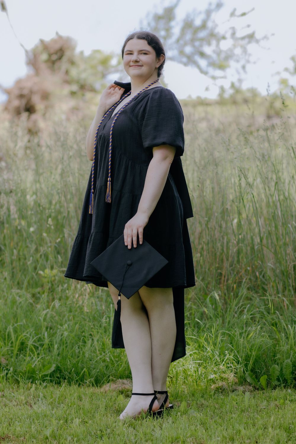 Graduate poses in black graduation gown against natural outdoor backdrop.