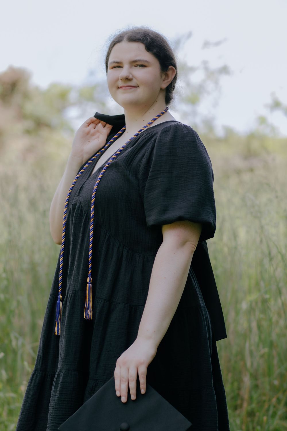 Graduate in black graduation dress holds cap while posing in meadow setting.