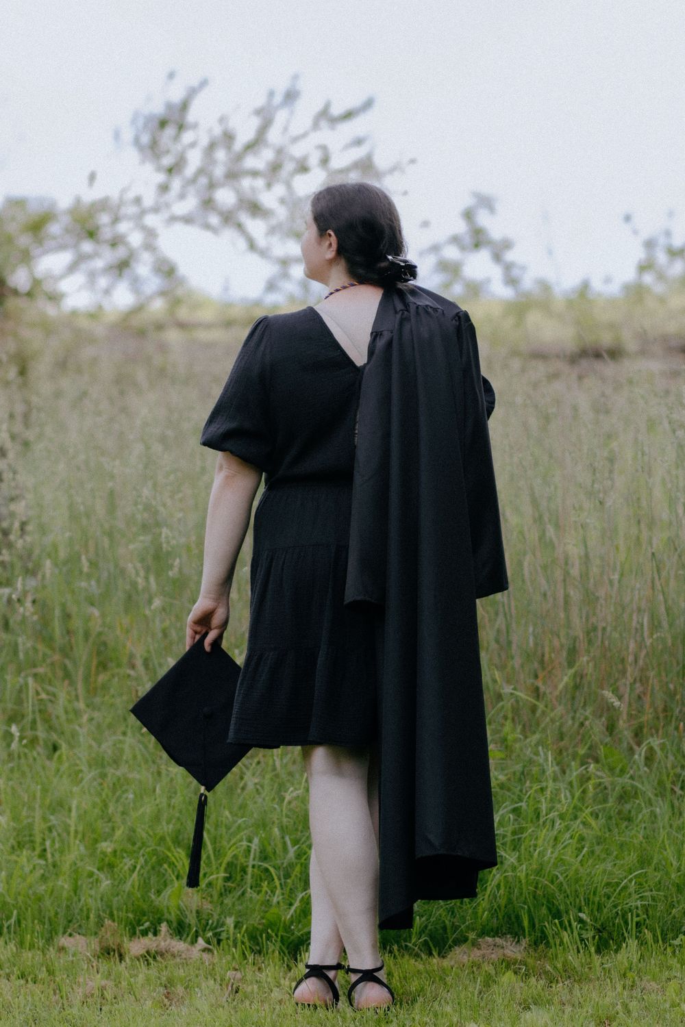 Graduate in black gown shown from behind holding cap in grassy field.