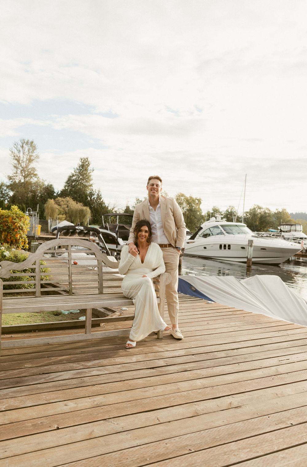 A couple poses together on a wooden dock with boats and a marina in the background during sunset.