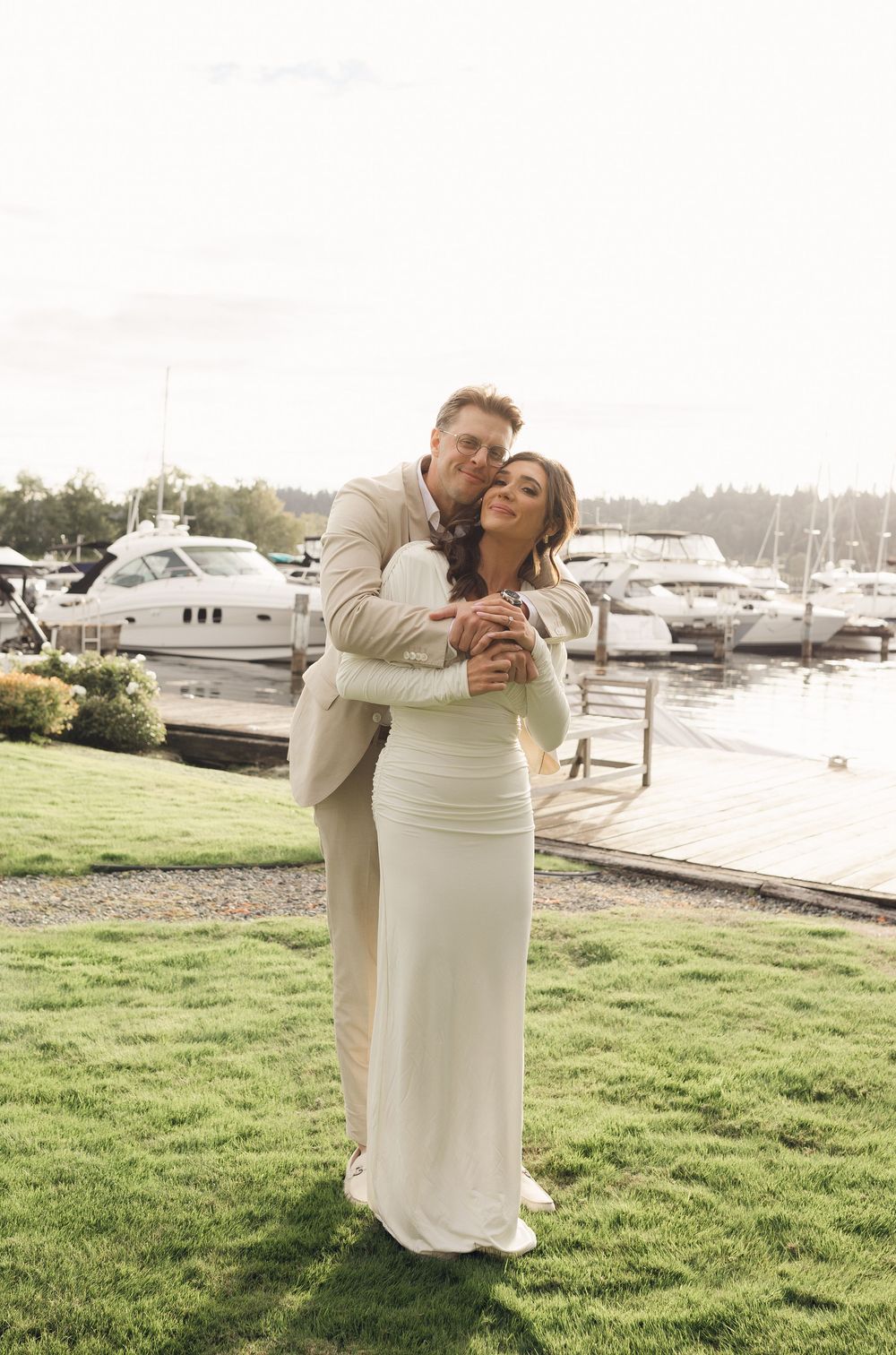 A couple in white and beige attire share romantic moments on a grassy lawn near a marina with boats in the background.