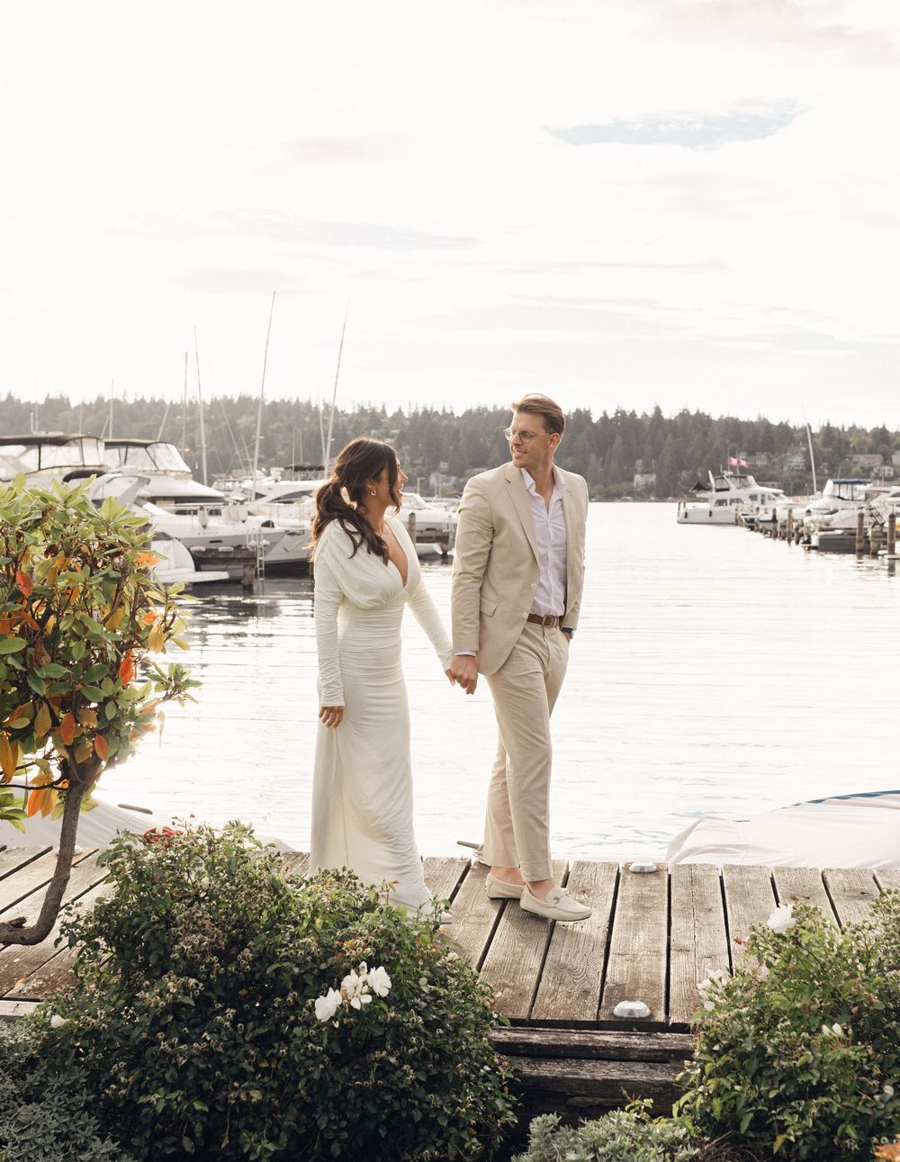 Couple in elegant white and beige attire walking along a wooden dock by a marina with boats and trees in background.