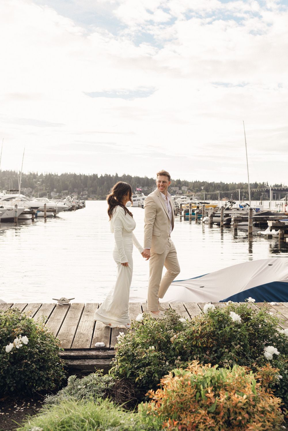 A couple in wedding attire walks along a scenic marina boardwalk with boats and flowers in the background.