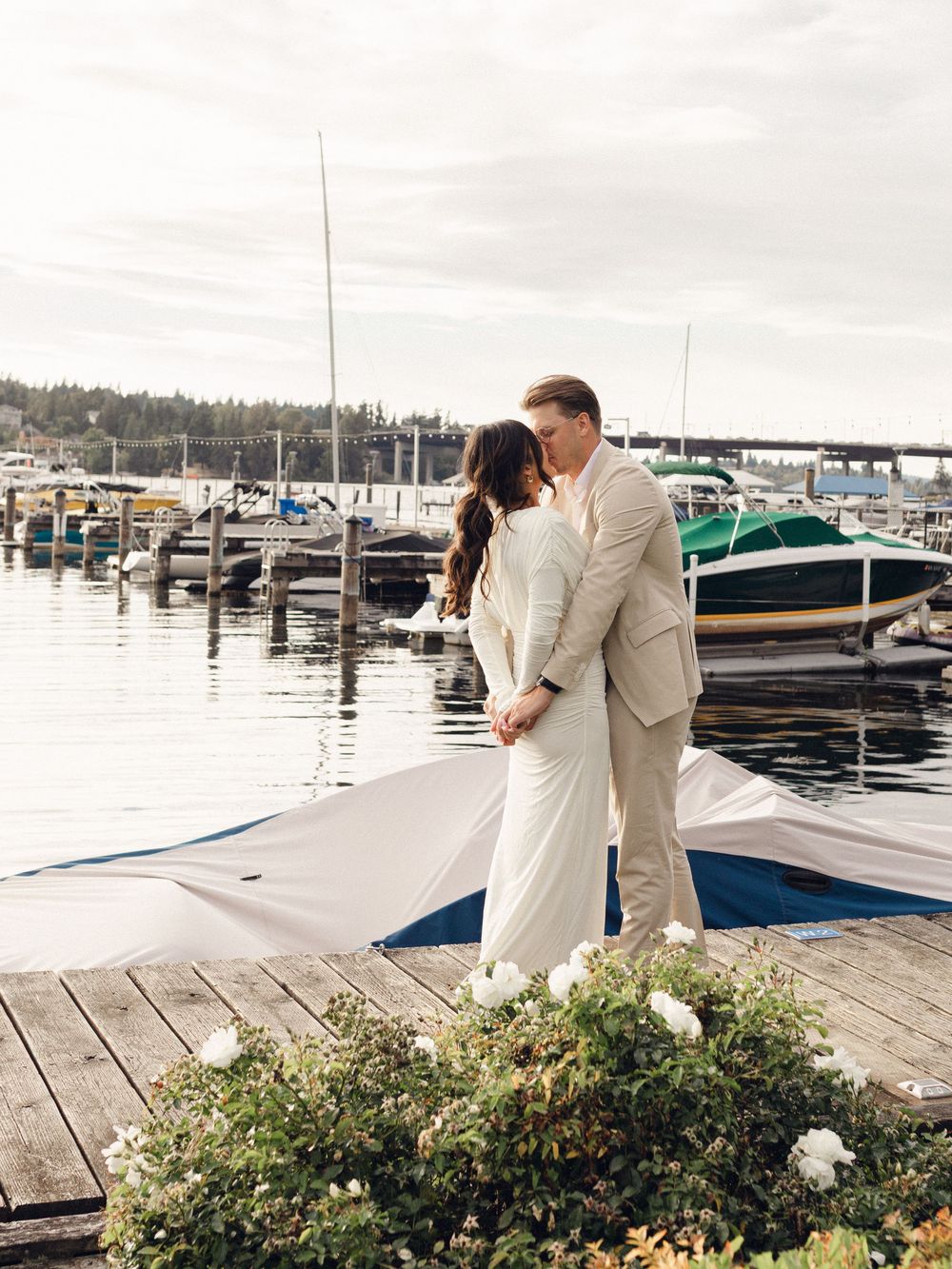 A couple shares a romantic moment on a wooden dock at a marina with boats and greenery in the background.