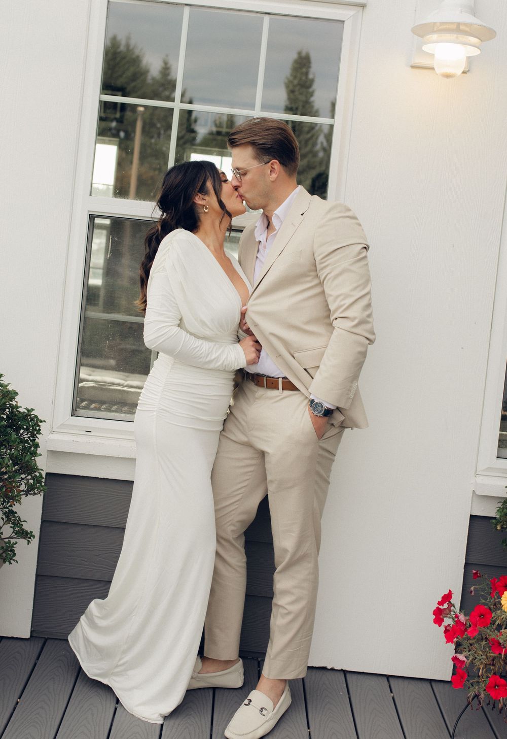 A romantic moment captured between newlyweds against a white exterior wall with red flowers.