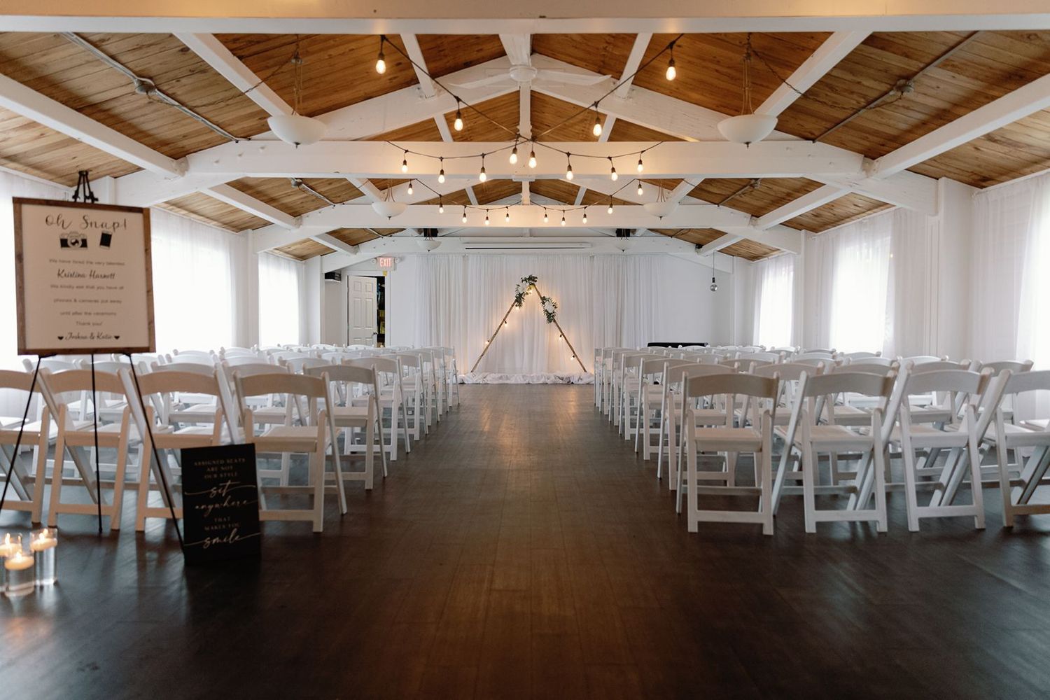 Bright white wedding venue interior with rows of white chairs, string lights, and a floral arch altar.