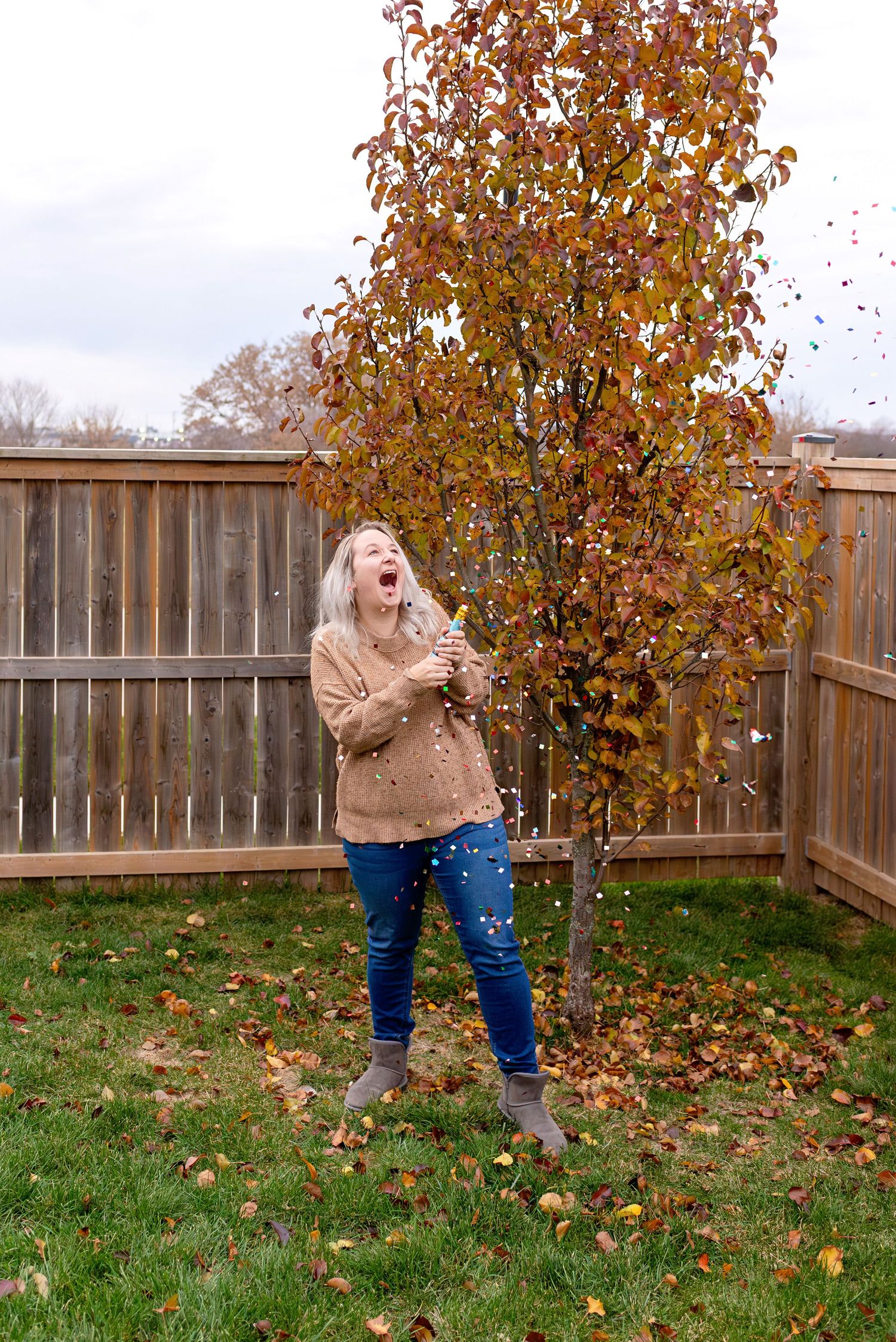Someone in a tan sweater and jeans enjoys a playful autumn moment with falling leaves in a backyard setting.