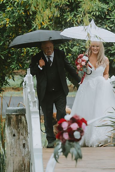 Two people walking with umbrellas at an outdoor wedding ceremony with red and white floral bouquets.