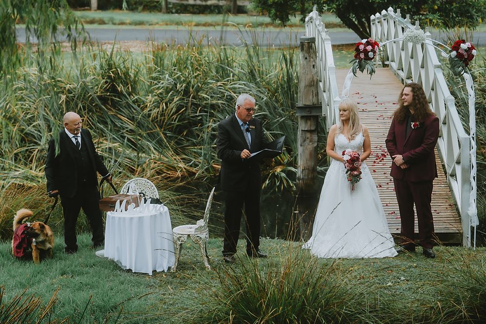 A wedding ceremony takes place in a lush garden setting with white picket fences and natural greenery.