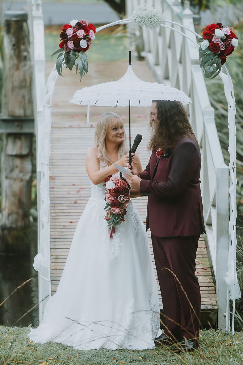 A romantic outdoor wedding scene with burgundy and white floral arrangements under a white draped archway.