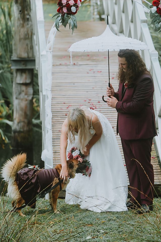 A bride in a white dress bends down to pet a dog while someone holds a white umbrella on a wooden walkway.
