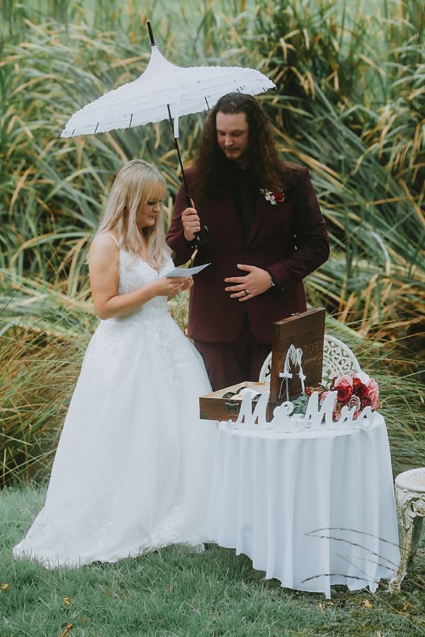 A couple shares an intimate moment at a white-clothed table outdoors with tall grass in the background.